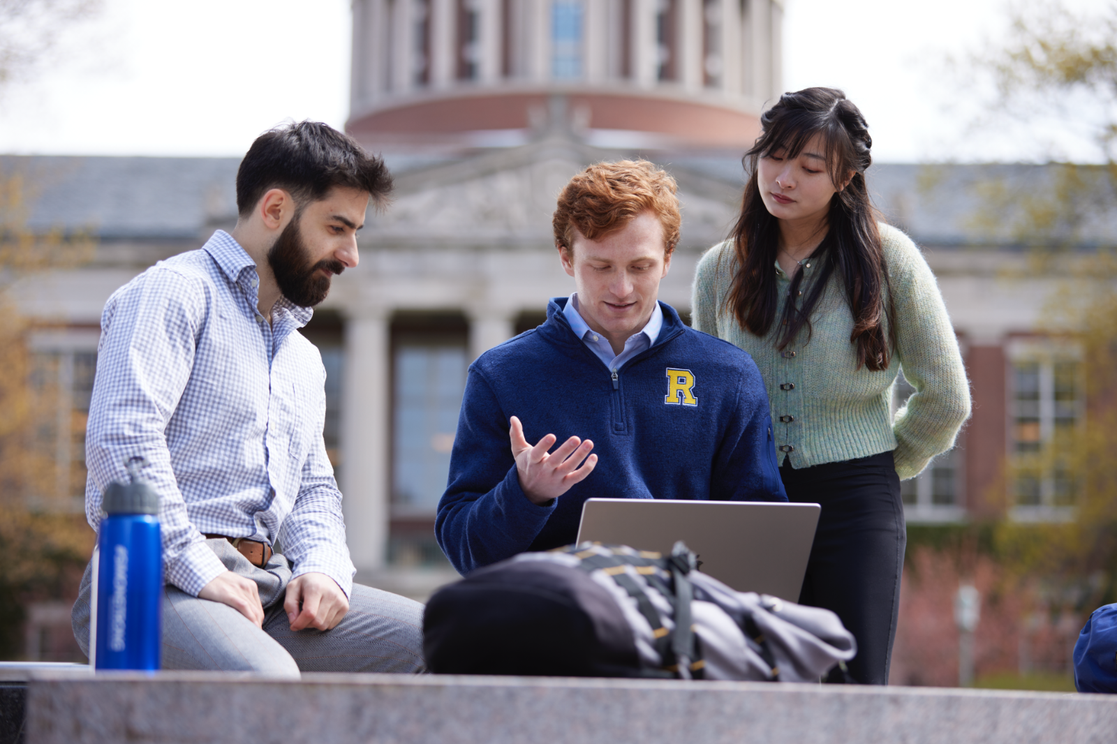 Three University of Rochester students sitting on a bench outside a building, engaged in conversation and enjoying the day.