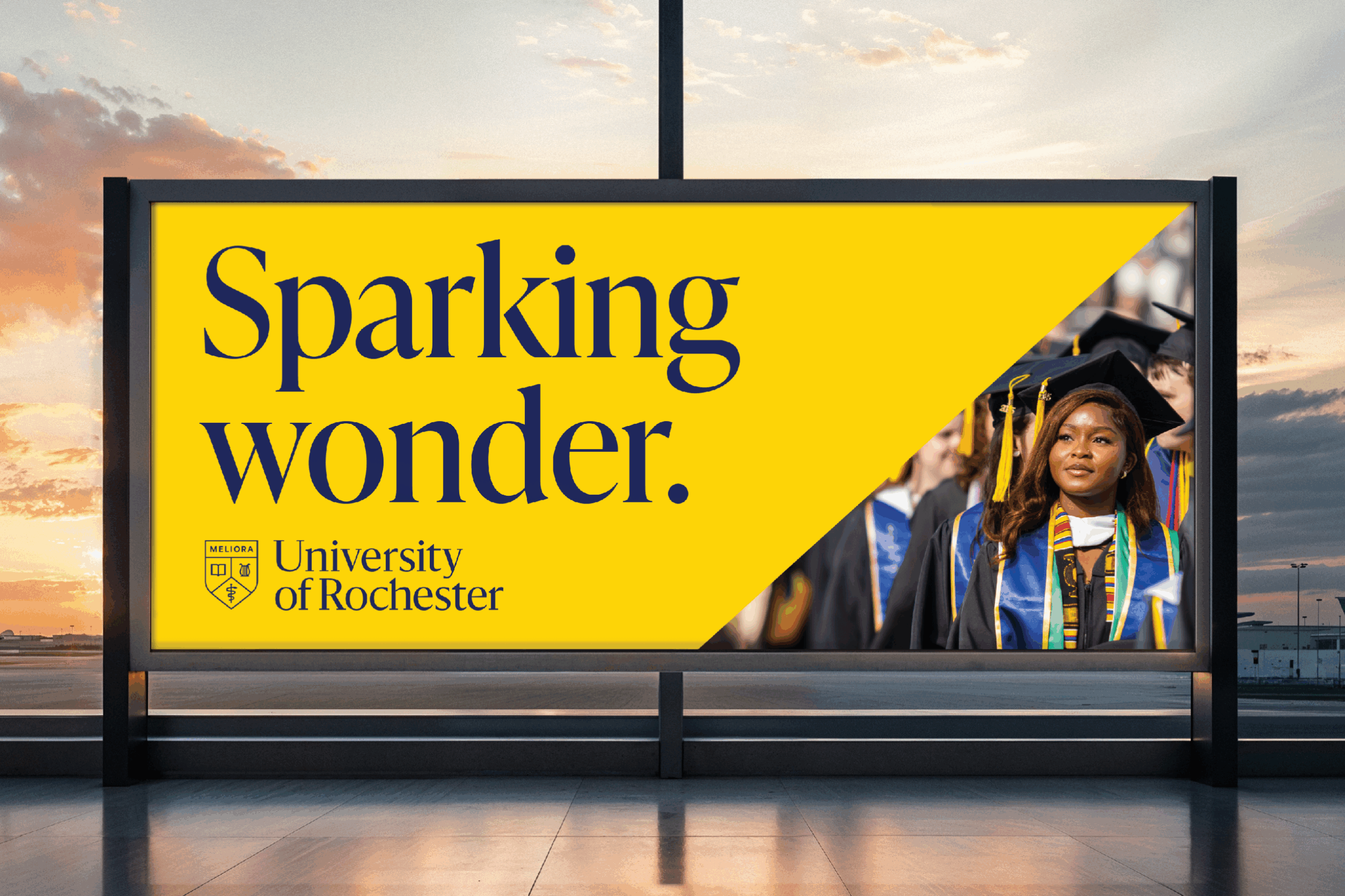 Woman in a graduation cap next to a sign that says sparking wonder and the university of rochester logo.